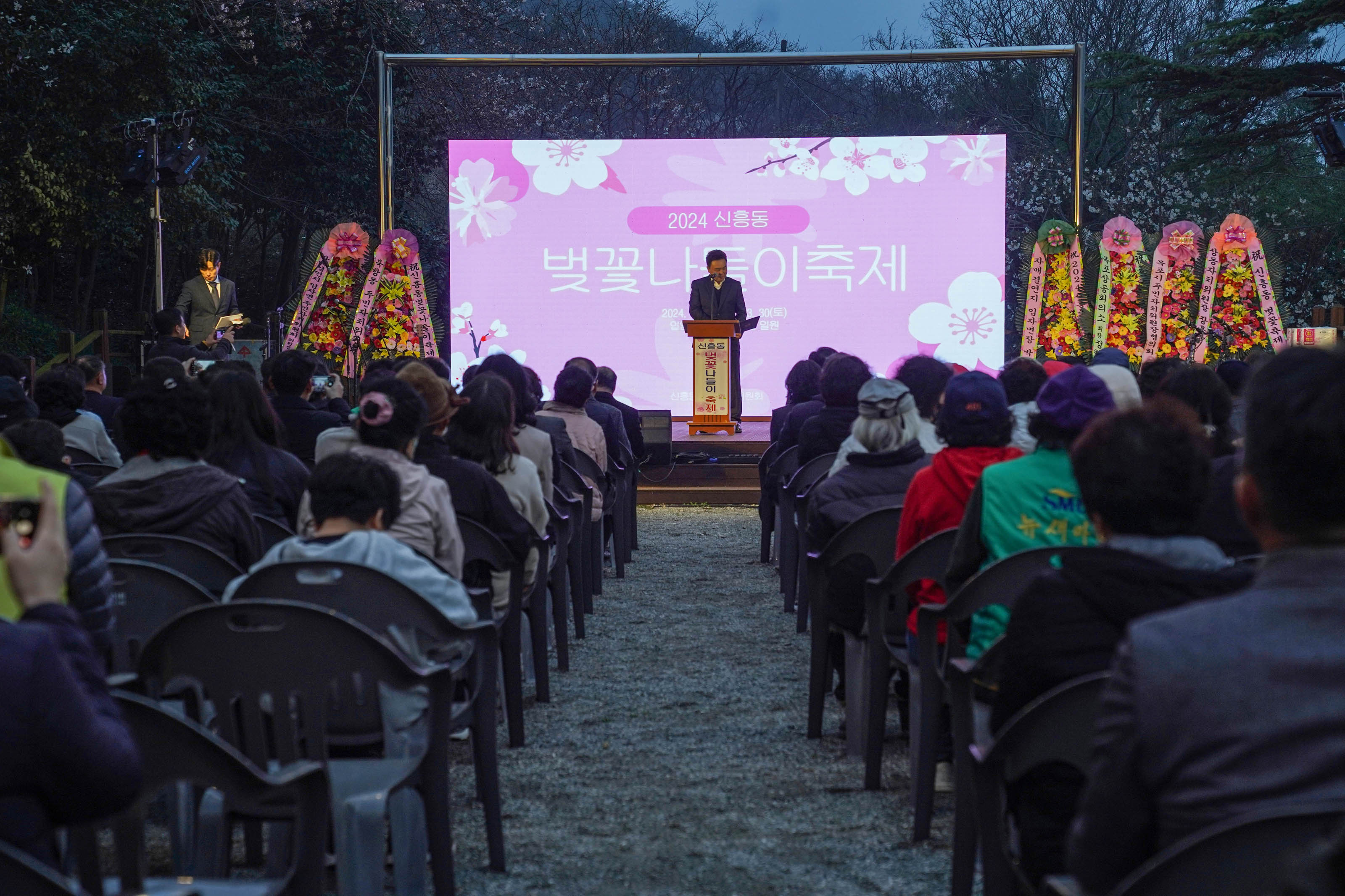 2024 신촌동 벚꽃나들이축제 화면이 핑크빛으로 띄워져 있고, 단상에선 중년의 남성. 6개의 화환도 보인다. 플라스틱 의자에 앉아 있는 축제에 참여한 시민들의 모습
