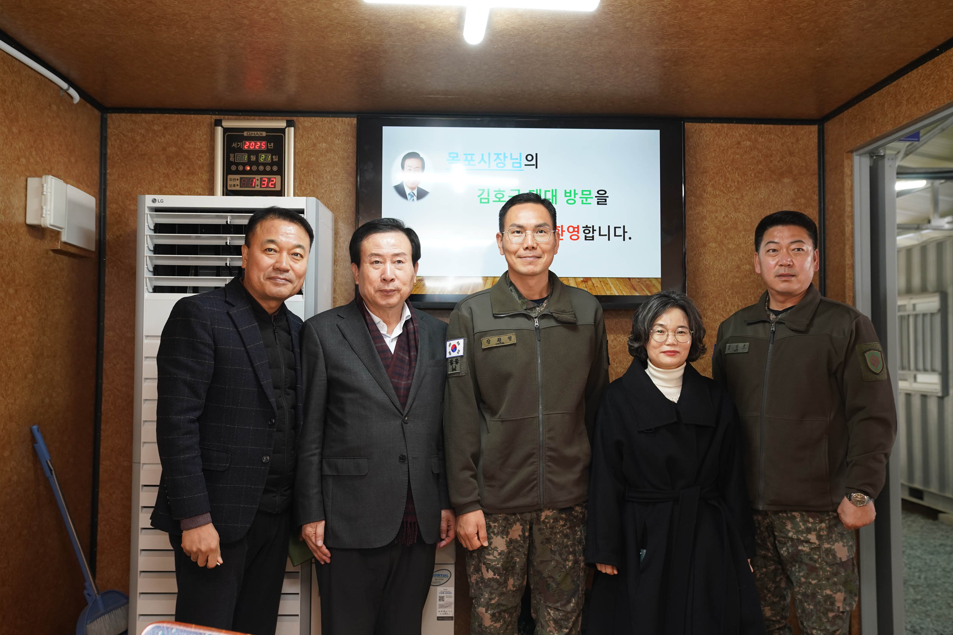 박홍률 목포시장이 군대 부대를 방문하여 관계자들과 기념 촬영을 하고 있다. 목포 시장 왼편에는 정장차림의 남성이 오른편에는 군복을 입은 군인2명과 검은색 외투를 입은 여성관계자가 있다. 배경에는 '목포시장님의 김호규 대대 방문을 환영합니다'라는 환영 메시지가 보인다. 