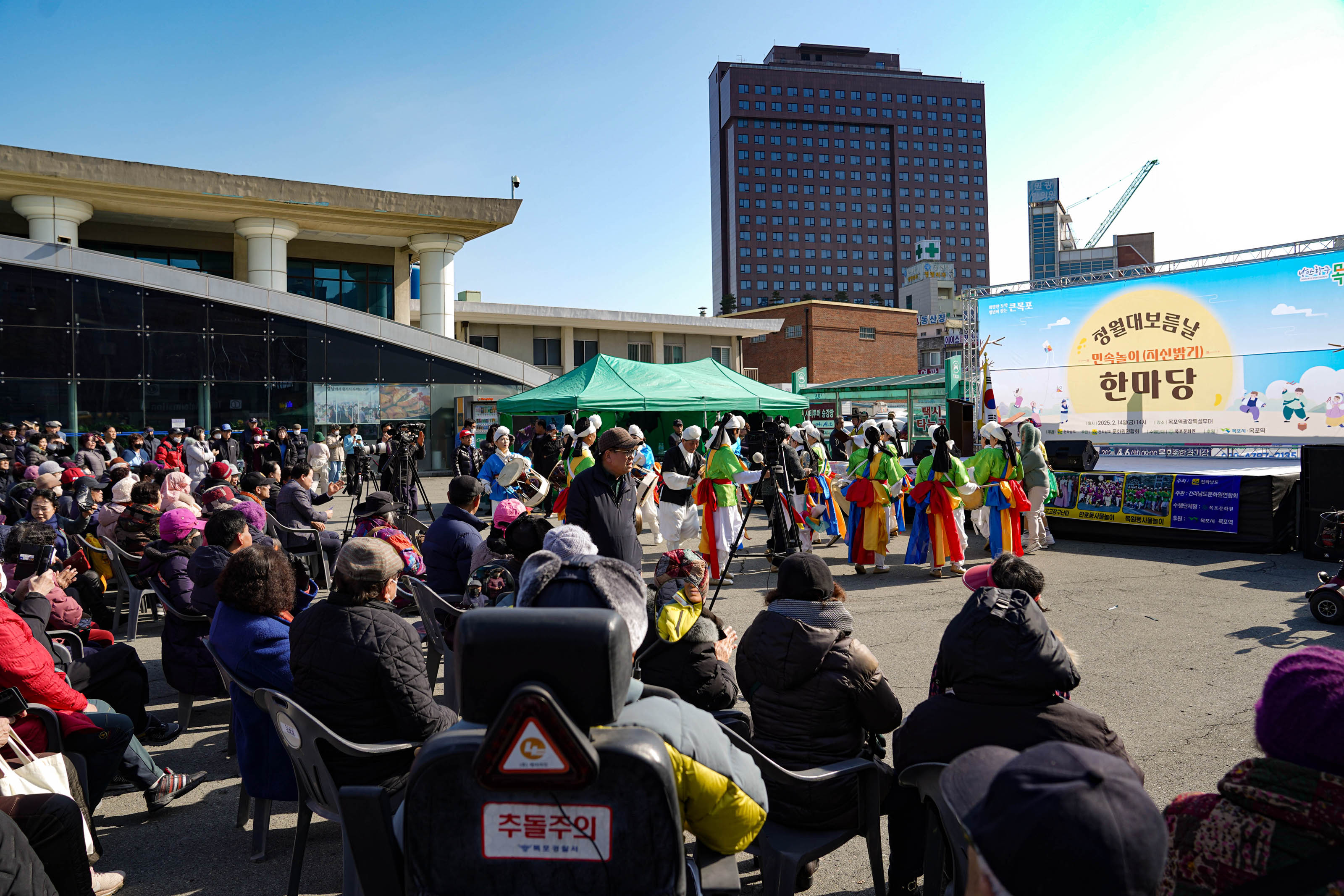 전통 복장을 갖춘 사물놀이 공연단이 무대 앞에서 연주를 준비하고 있으며, 객석에는 다양한 연령층의 시민들이 앉아 관람 중임. 무대에는 ‘청월대보름날 민속놀이(지신밟기) 한마당’이라는 문구가 적힌 배너가 걸려 있음. 무대 오른쪽에는 대형 꽃바구니가 놓여 있고, 무대 왼쪽에는 녹색 천막이 설치되어 있음. 무대 위 배너에는 ‘낭만항구 목포’ 문구가 보임.