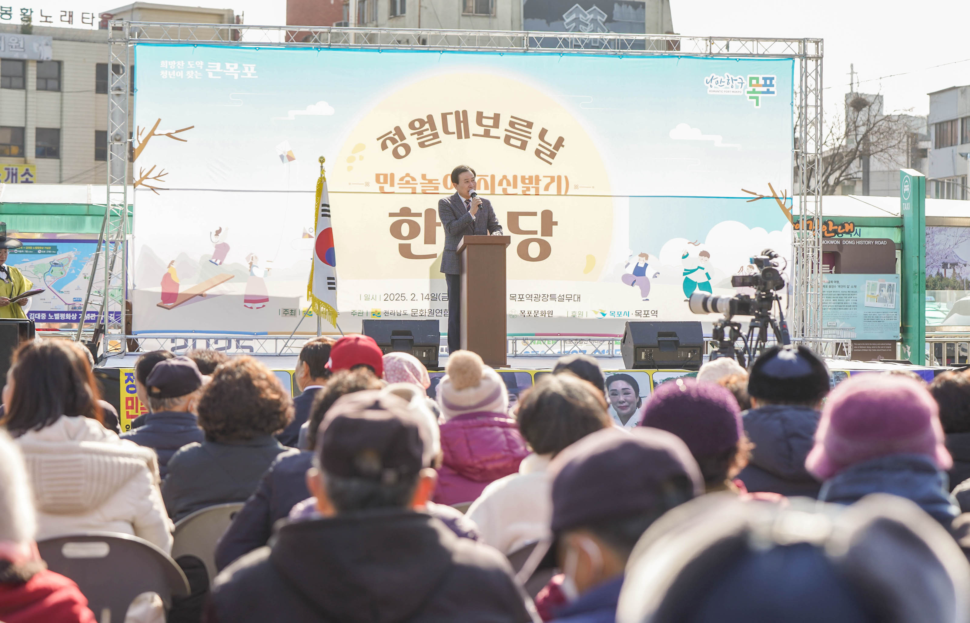 박홍률 목포시장이 무대에서 마이크를 들고 개회사를 하고 있으며, 청중들은 무대를 바라보고 있음. 무대에는 ‘청월대보름날 민속놀이(지신밟기) 한마당’이라고 적힌 대형 배너가 걸려 있고, 좌측 상단에는 ‘희망찬 도약 청년이 찾는 큰 목포’와 ‘낭만항구 목포’ 문구가 보임. 행사 일시는 2025년 2월 14일 금요일 오후 2시로 표기되어 있고, 장소는 목포역광장 특설무대임.