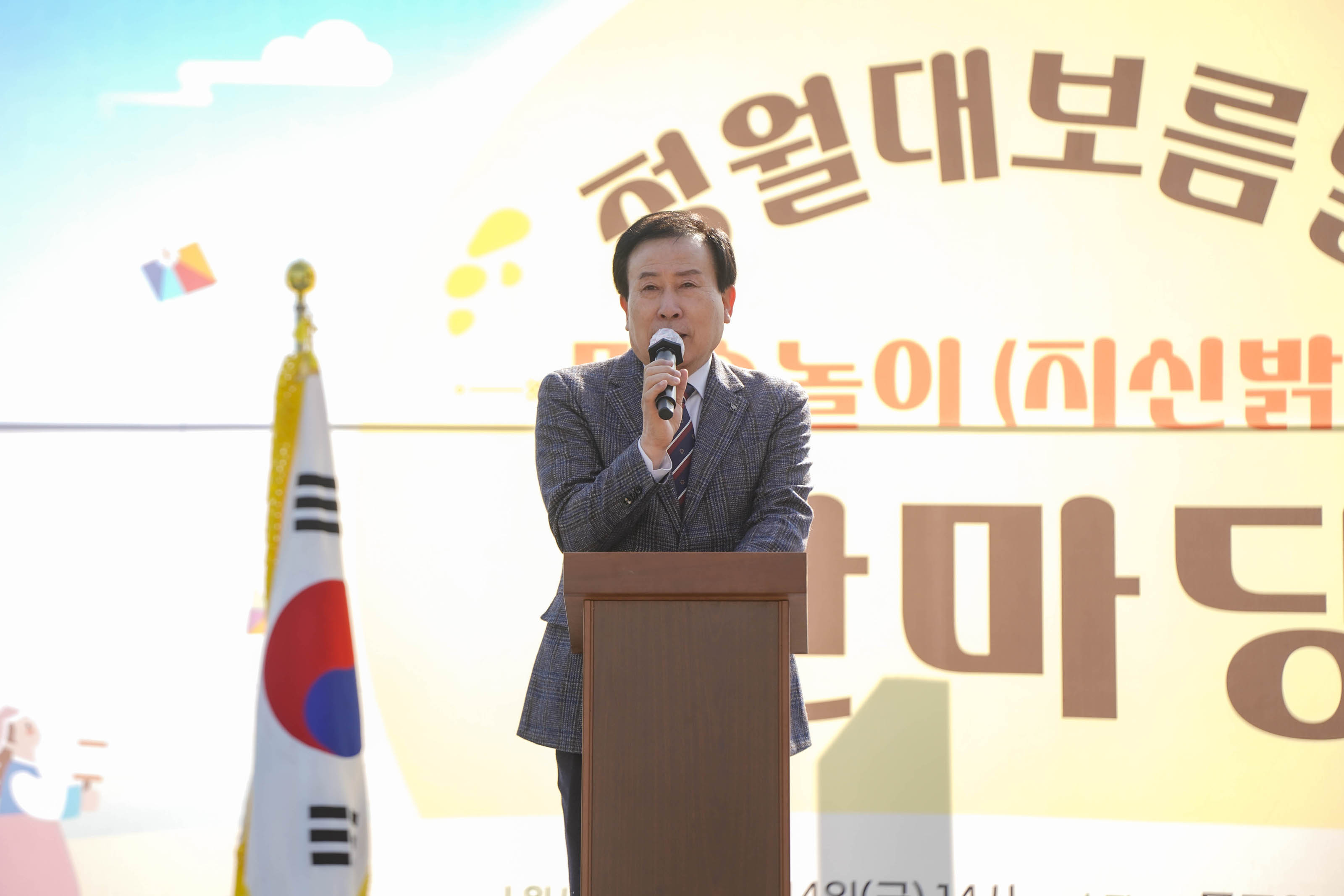 무대 중앙에서 정장을 입은 박홍률 목포시장이 마이크를 들고 연설 중이며, 배경에는 ‘청월대보름날 민속놀이(지신밟기) 한마당’ 문구가 큼지막하게 보임. 무대 왼쪽에는 태극기가 세워져 있음.
