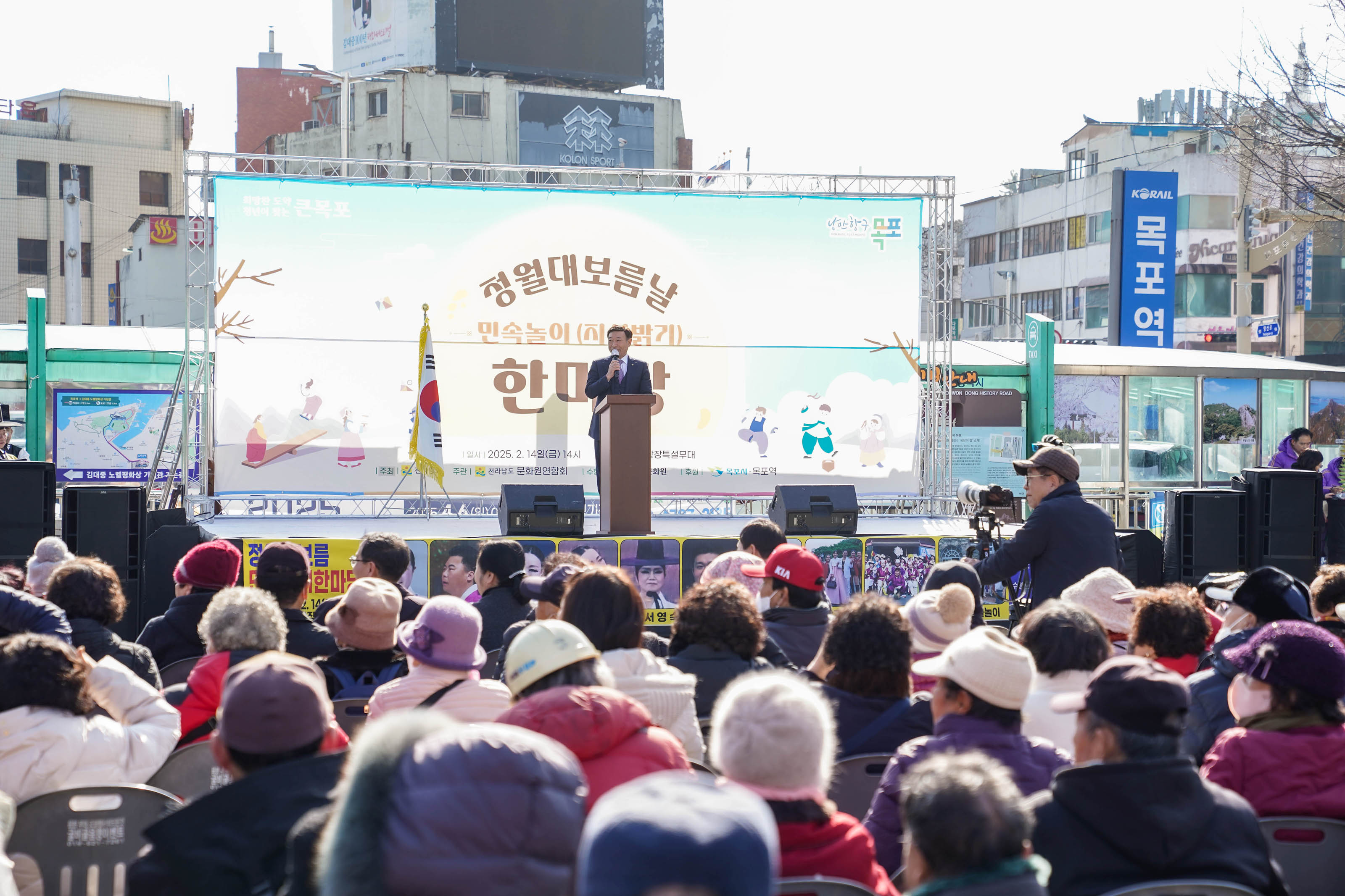 청중들이 무대를 바라보고 있으며, 무대에는 관계자가 마이크를 들고 연설 중임. 배경 배너에는 ‘청월대보름날 민속놀이(지신밟기) 한마당’이라는 문구가 적혀 있고, 태극기가 함께 배치되어 있음. 현수막 하단에 ‘2025. 2. 14(금) 14시 목포역광장특설무대’와 ‘전라남도, 문화관광협회, 목포문화원, 목포시, 목포역’ 문구가 명시되어 있음.