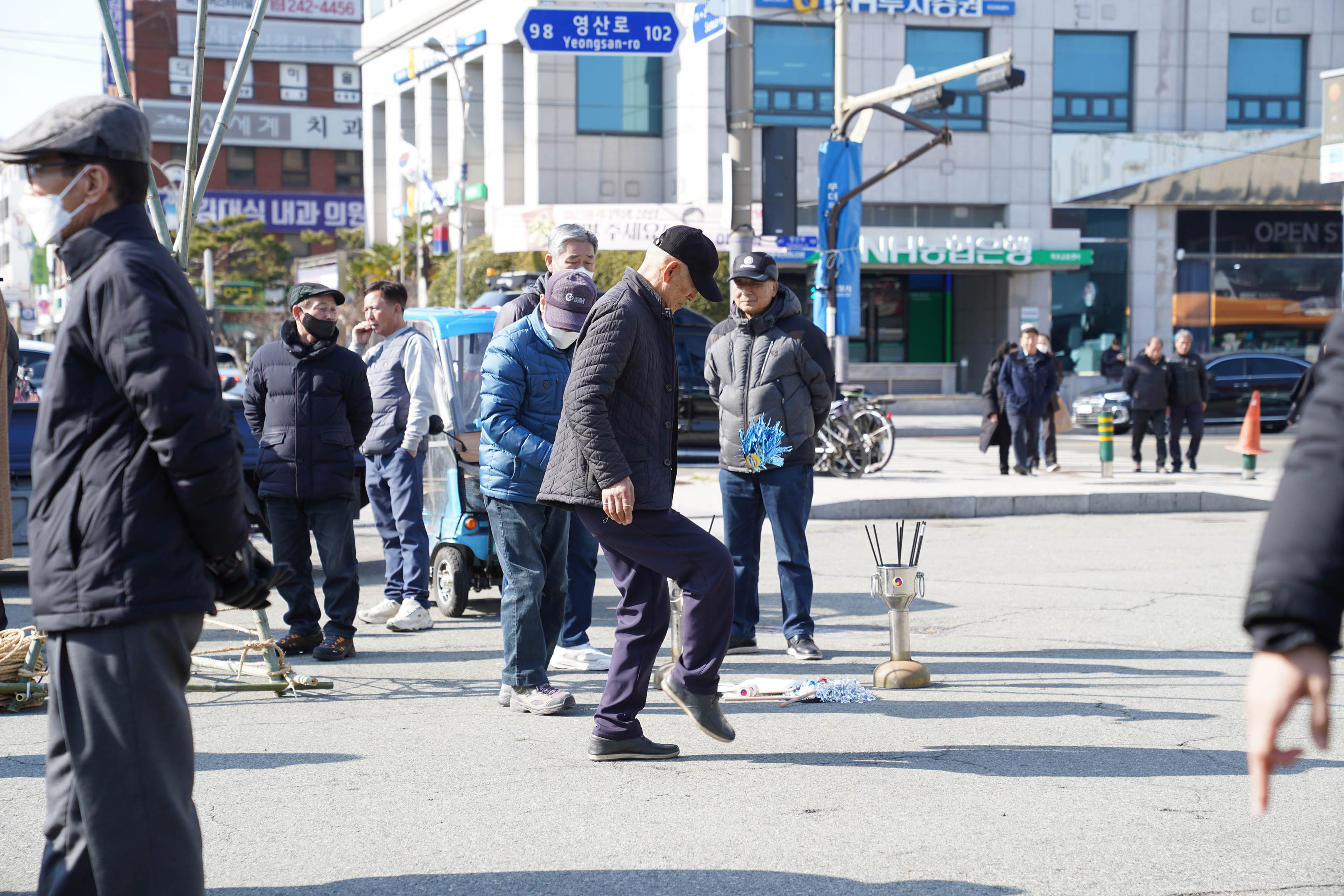 중장년 남성이 발로 공중에 있는 고무신을 차며 민속놀이를 하고 있고, 주변에는 여러 시민들이 서서 지켜보고 있음. 배경에는 ‘영산로’, ‘NH농협은행’, ‘OPEN SPACE’ 등의 간판이 보이고, 길가에는 윷놀이 도구와 장애물 등이 놓여 있음.