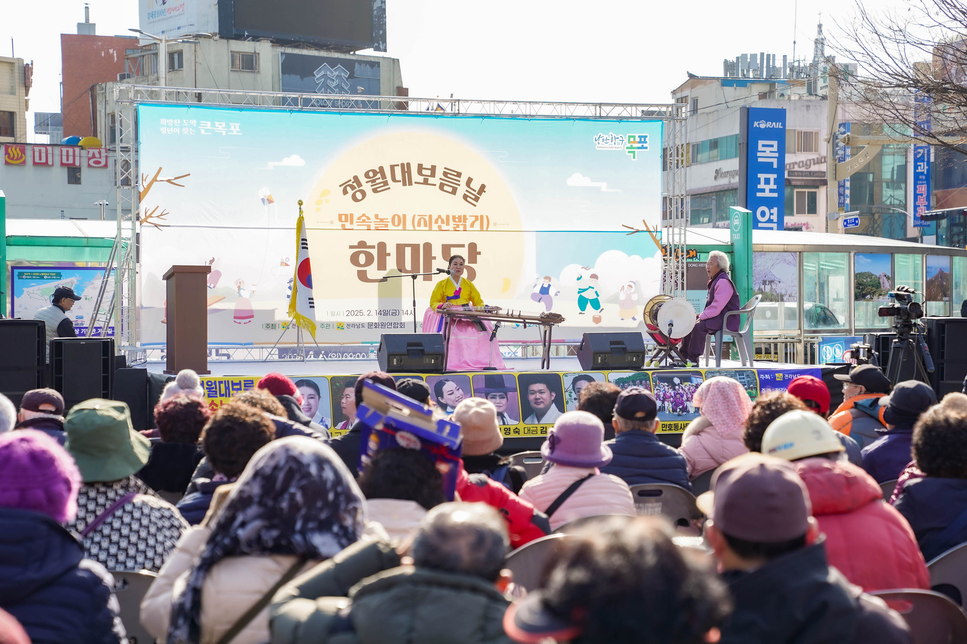 무대 위에서 한복을 입은 국악인이 가야금을 연주하고, 옆에 장구 연주자가 함께 연주 중임. 무대 배경에는 ‘청월대보름날 민속놀이(지신밟기) 한마당’이라는 대형 배너가 있으며, 행사 주최 및 후원(전라남도, 문화관광협회, 목포문화원, 목포시, 목포역)이 표기되어 있음. 무대 하단에는 지역 예술인 사진들이 나열되어 있고, 행사 진행 현장이 시민들로 가득 차 있음.