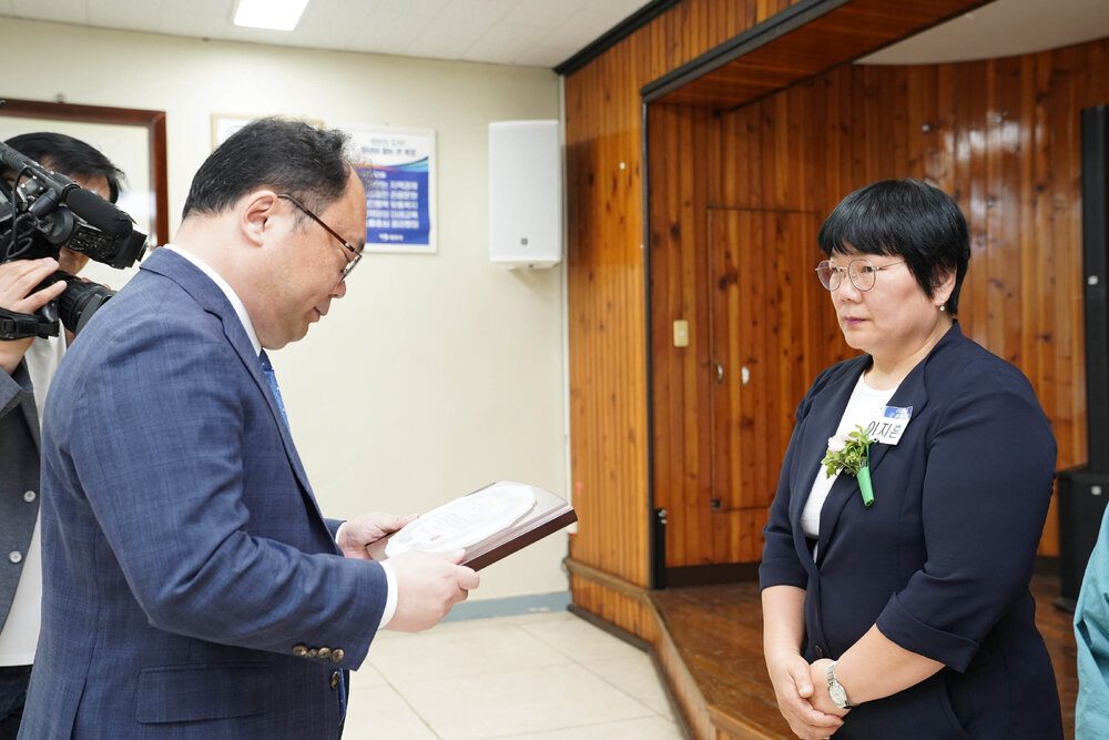 정장을 입고 안경을 낀 남성이 검은색 7부 자켓을 입은 여성에게 패를 수여하고 있다
