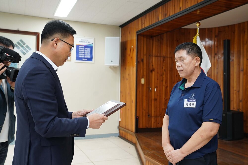 정장 차림의 관계자가 남색 반팔 티셔츠를 입은 남성 수상자에게 패를 전달하며 읽고 있는 모습이다.