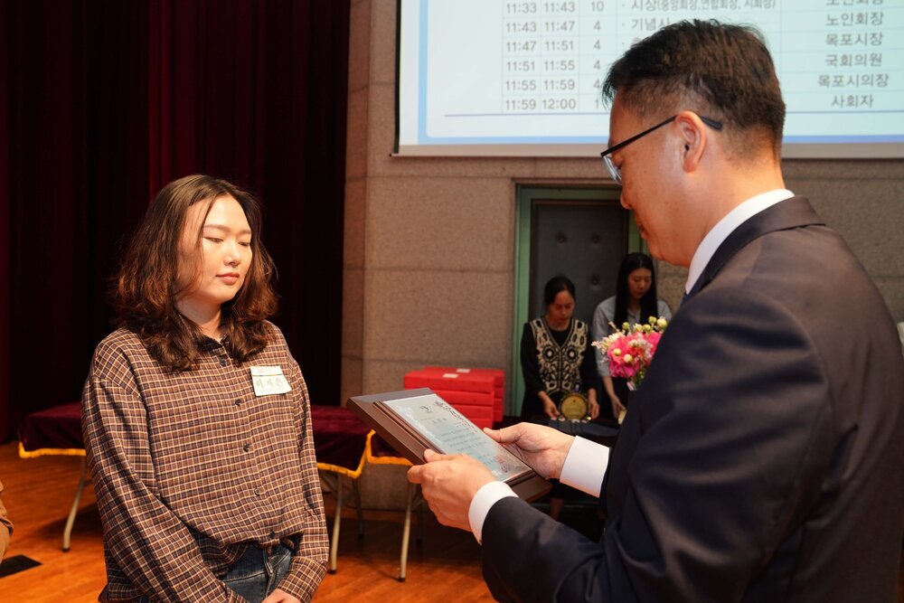 정장 차림의 관계자가 체크무늬 상의를 입은 여성 수상자에게 패를 전달하며 읽고 있는 모습이다.