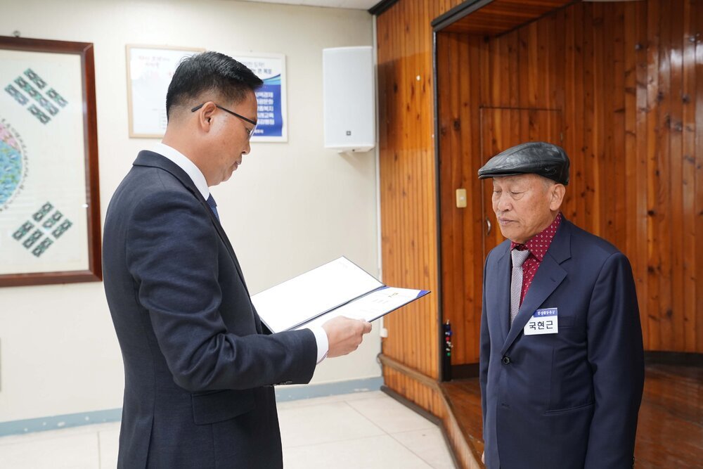11월 정례조회로 정장을 입고 안경을 쓴 남성이 머리에 모자를 쓰고 짙은 네이비색 정장을 입은 국현근 수상자에게 상장을 수여 하고 있다.