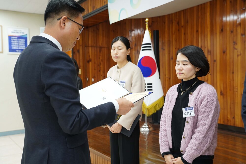 11월 정례조회로 정장을 입고 안경을 쓴 남성이 검은색 티에 연보라색 가디건을 입은 유경화 수상자에게 상장을 수여 하고 있다.