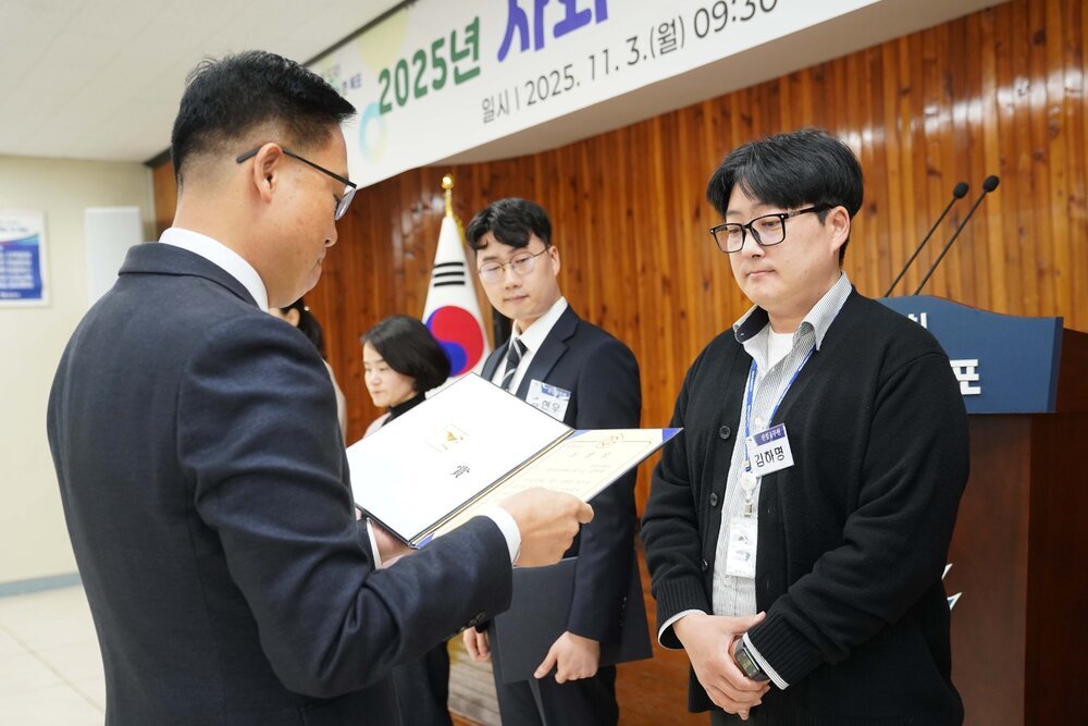 11월 정례조회로 정장을 입고 안경을 쓴 남성이 안경을 쓰고 흰색 셔츠에 검은색 가디건을 입고 사원증을 착용한 채 두 손을 모은 김하명 수상자에게 상장을 수여 하고 있다.
