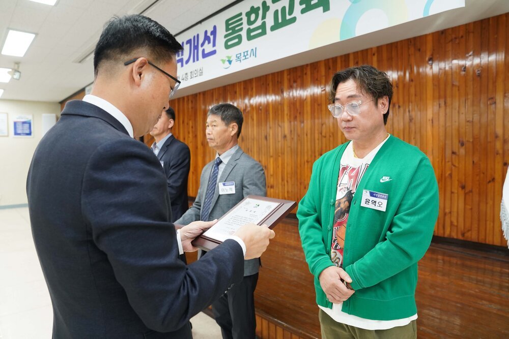 11월 정례조회로 정장을 입고 안경을 쓴 남성이 곱슬머리에 흰색 안경을 쓰고 초록색 가디건을 입은 윤역오 수상자에게 상패를 수여 하고 있다.