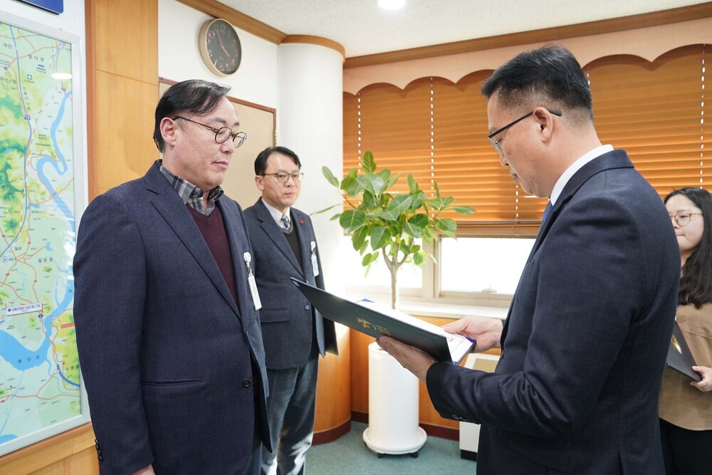 2026년 상반기 4급이하 정기인사로 정장을 입은 남성이 실내 사무실에서 한 남성에게 상장을 전달하며 내용을 읽고 있고, 남성은 안경을 쓰고 남색 자켓에 와인색 니트를 겹쳐 입은 채 사원증을 착용하고 서 있으며 왼쪽 벽면에는 대형 지도가 걸려 있다.