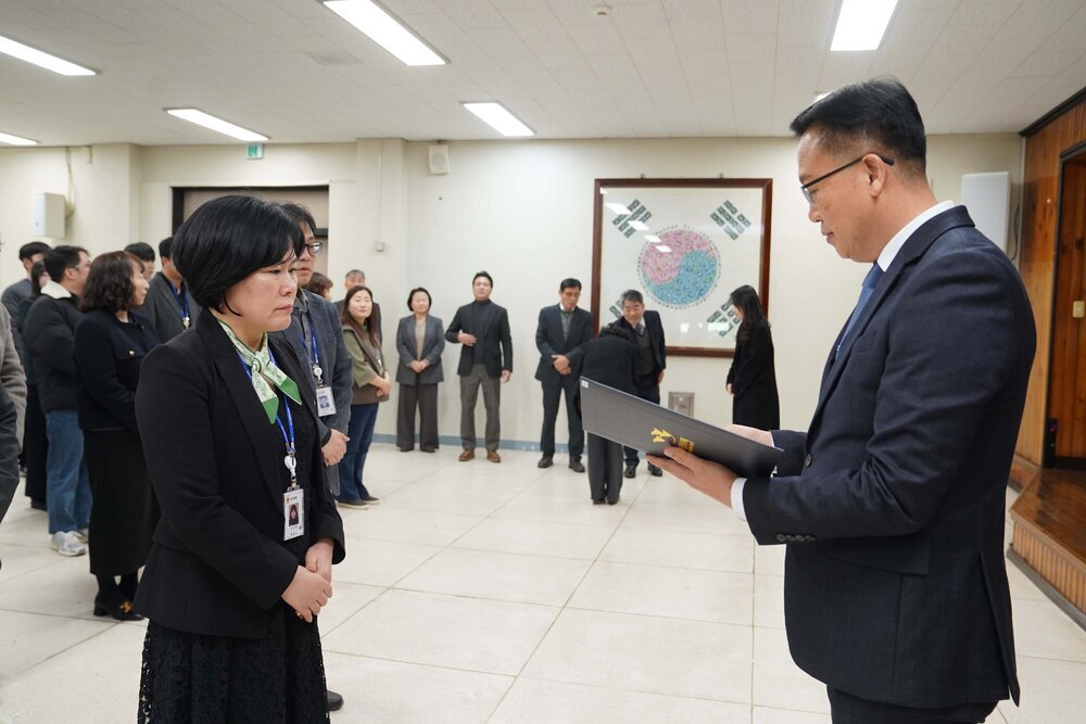 2026년 상반기 4급이하 정기인사로 정장을 입은 남성이 실내 행사장에서 한 여성에게 상장을 전달하며 내용을 읽고 있고, 여성은 검은색 자켓에 초록색 무늬가 있는 스카프를 매고 사원증을 착용한 채 두 손을 모은 상태로 서 있으며 뒤쪽에는 참석자들이 행사를 지켜보고 있다.