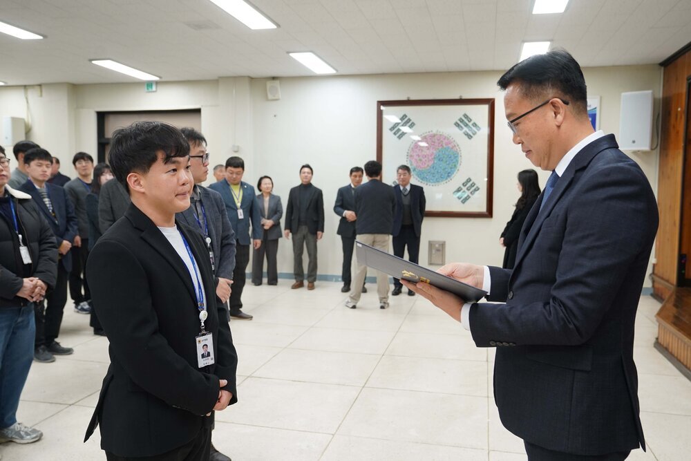 2026년 상반기 4급이하 정기인사로 정장을 입은 남성이 실내 행사장에서 한 남성에게 상장을 전달하며 내용을 읽고 있고, 남성은 검은색 정장 자켓을 입고 사원증을 착용한 채 두 손을 모은 상태로 서 있으며 뒤쪽으로 참석자들이 줄지어 서서 행사를 지켜보고 있다.