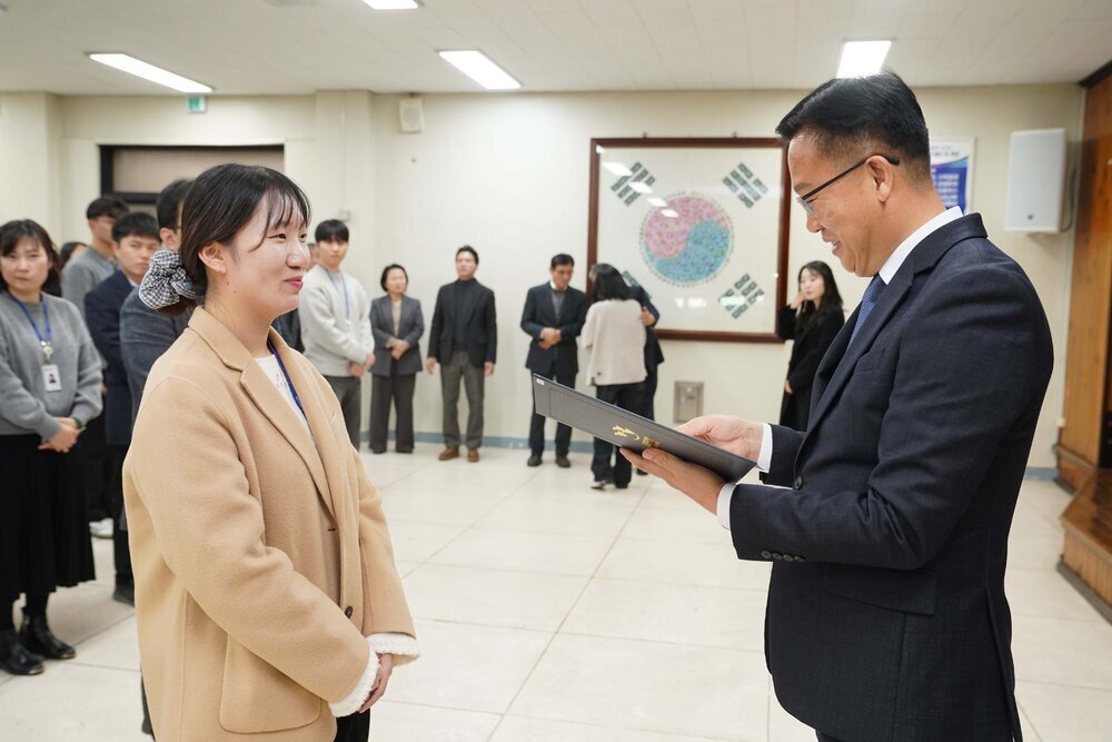 2026년 상반기 4급이하 정기인사로 정장을 입은 남성이 실내 행사장에서 한 여성에게 상장을 전달하며 내용을 읽고 있고, 여성은 체크무늬 머리끈으로 머리를 묶고 밝은 베이지색 코트를 입고 사원증을 착용한 채 두 손을 모은 상태로 서 있으며 뒤쪽으로 참석자들이 줄지어 서서 행사를 지켜보고 있다.