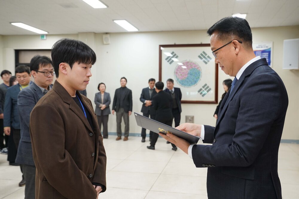 2026년 상반기 4급이하 정기인사로 정장을 입은 남성이 실내 행사장에서 한 남성에게 상장을 전달하며 내용을 읽고 있고, 남성은 갈색 정장 자켓을 입고 두 손을 모은 상태로 서 있으며 뒤쪽으로 참석자들이 줄지어 서서 행사를 지켜보고 있다.
