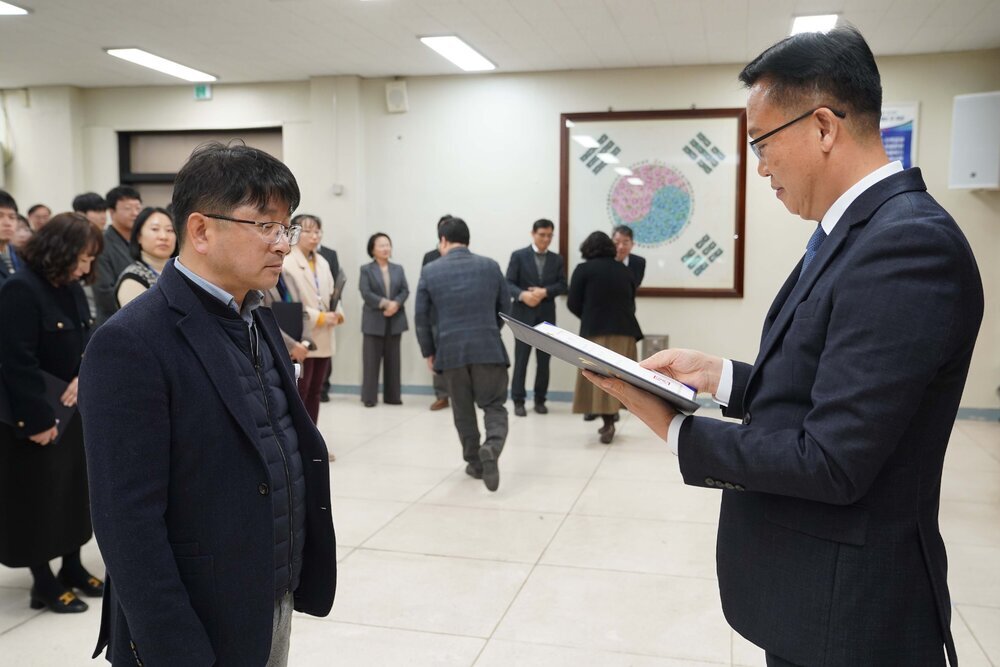 2026년 상반기 4급이하 정기인사로 정장을 입고 안경을 쓴 남성이 실내 행사장에서 한 남성에게 상장을 전달하며 내용을 읽고 있고, 남성은 안경을 쓰고 어두운 자켓을 입은 채 서 있으며 뒤쪽에는 여러 사람들이 줄지어 서서 행사를 지켜보고 있다.