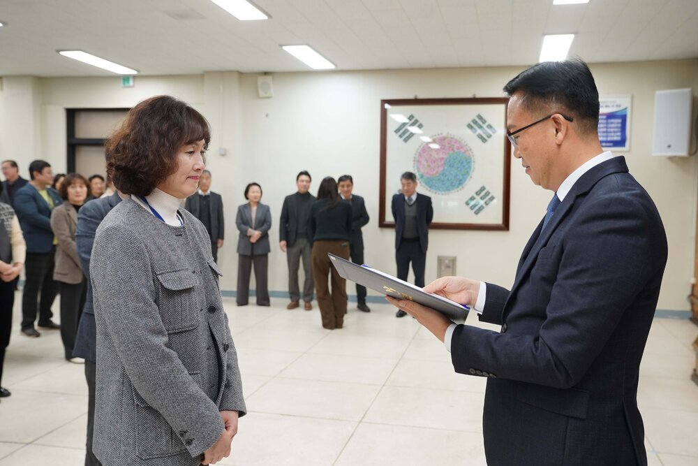 2026년 상반기 4급이하 정기인사로 정장을 입고 안경을 쓴 남성이 실내 행사장에서 한 여성에게 상장을 전달하며 내용을 읽고 있고, 여성은 회색 자켓을 입고 두 손을 모은 채 서 있으며 뒤쪽에는 여러 사람들이 줄지어 서서 행사를 지켜보고 있다.