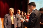 정장 차림의 관계자가 주황색 셔츠와 재킷을 입은 남성 수상자에게 패를 전달하며 읽고 있는 모습이다.