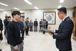 2026년 상반기 4급이하 정기인사로 정장을 입은 남성이 실내 행사장에서 한 남성에게 상장을 전달하며 내용을 읽고 있고, 남성은 검은색 맨투맨 티셔츠를 입고 사원증을 착용한 채 두 손을 내린 상태로 서 있으며 뒤쪽으로 참석자들이 줄지어 서서 행사를 지켜보고 있다.